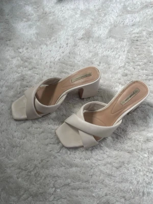 Vita slip-in sandaler med klack - Säljer ett par vita slip-in sandaler från Stradivarius med breda korslagda remmar och fyrkantig tå. Klackarna är blockformade och ger en trendig look. Tillverkade i konstläder med mjuk innersula för extra komfort. I använt skick, se bilder för att se skicket. 
