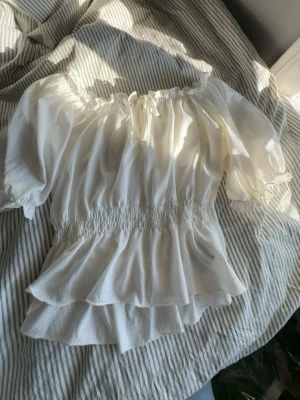 Vit offshoulder blus med volang - Söt vit offshoulder blus med volangdetaljer och resår i midjan. Blusen har trekvartsärm och en liten rosett framtill vid halsen. Tillverkad i ett lätt, luftigt tyg som ger en romantisk vibe.