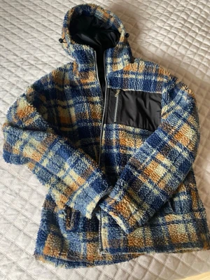 Rutig teddyjacka med huva - Mysig teddyjacka i blått, gult och orange rutmönster. Jackan har huva, dragkedja framtill, svart bröstficka med dragkedja och två sidofickor. Fodrad insida i svart tyg. Perfekt för kalla dagar och ger en cool retrovibe.