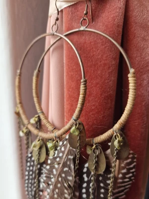 Stora boho örhängen med fjädrar hippie bohem - Unika stora örhängen i boho-stil med bruna och vita fjädrar, guldfärgade kedjor och små metallbrickor samt stenar. Ringen är lindad med beige tråd för en naturlig känsla. Perfekta för dig som vill sticka ut med en cool och kreativ accessoar. Handgjorda och köpta I Costa Rica. Använda ändast några gånger. 