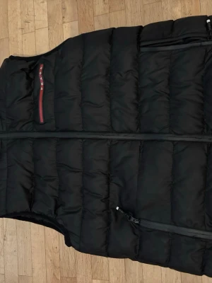 Svart dunväst från Prada - Snygg svart dunväst från Prada med quiltad design och hög krage. Västen har två dragkedjeförsedda fickor framtill och en diskret logga på bröstet med röd detalj. Perfekt för lager-på-lager och streetstyle.