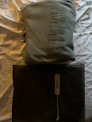 Grön Essentials Fear of God hoodie - Snygg grön hoodie från Essentials Fear of God med stor logga framtill som jag säljer eftersom den är lite liten för mig. Klassisk passform med huva och mjukt material som känns riktigt skönt.