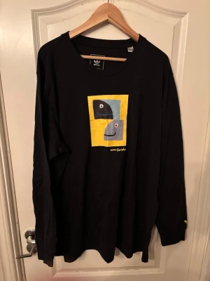 Adidas skateboarding shmoofoil Svart långärmad topp  - Svart långärmad topp från Adidas med färgglad grafisk print framtill i gult, blått och svart. Rund halsringning och relaxed fit. Perfekt för dig som gillar streetwear och vill ha något unikt i garderoben.