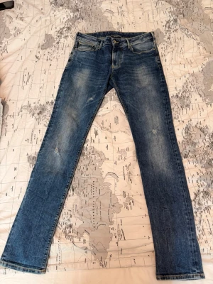 Blå raka jeans W29 L32 - Säljer ett par klassiska blå jeans med raka ben och lätt slitna detaljer. Jeansen har fem fickor, gylf med dragkedja och knapp, samt kontrastsömmar. Perfekta för en avslappnad och trendig look.
