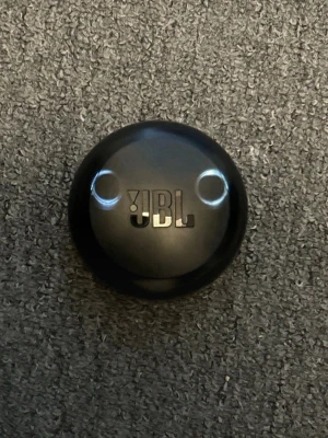 JBL svarta trådlösa in-ear hörlurar - Snygga svarta trådlösa in-ear hörlurar från JBL med rund laddningsetui. Hörlurarna har en modern design med JBL-logga på varje plugg och laddas smidigt i det kompakta fodralet. Perfekta för musik, poddar och samtal på språng.