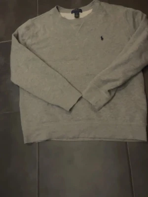 Grå stickad tröja från Polo Ralph Lauren - Klassisk grå stickad tröja från Polo Ralph Lauren med rund halsringning och diskret broderad logga på bröstet. Tröjan har långa ärmar och ribbade muddar vid ärmslut och nederkant. Perfekt för lager på lager och enkel att matcha.