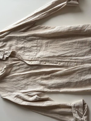 Beige linneskjorta från Zara - Säljer en stilren beige skjorta från Zara i mjukt linne. Skjortan har klassisk krage, bröstficka och knäppning framtill. Perfekt för en avslappnad och trendig look.