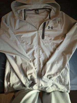 Ljusgrå vindjacka från Under Armour - Snygg ljusgrå vindjacka från Under Armour med huva och hel dragkedja framtill. Jackan har elastiska muddar, två sidofickor och en svart logga på bröstet. Materialet är tunt och smidigt, perfekt för blåsiga dagar. Diskret Under Armour-tryck på mudden.