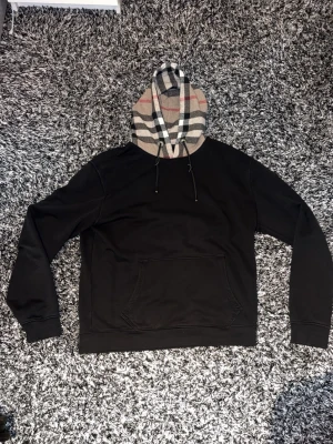 Burberry hoodie - Svart hoodie med klassisk Burberry-rutig huva i beige, svart, vitt och rött. Hoodien har dragsko med metallöglor och en stor magficka framtill. Pris går att diskutera 