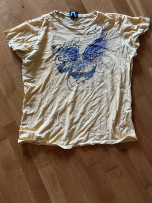 Gul Harley-Davidson t-shirt - Säljer en gul t-shirt från Harley-Davidson med tryck i svart och lila framtill. T-shirten har rund hals och korta ärmar. Materialet känns som bomull och trycket har coola detaljer med vingar och snirkliga mönster! Storlek är XL men mindre storlekar hade passat skitfint!!