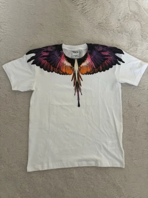 Vit t-shirt med färgglada vingar - Säljer en vit t-shirt från Marcelo Burlon med ikoniskt tryck av färgglada vingar i lila, orange och svart över axlar och bröst. T-shirten har rund hals och korta ärmar. Perfekt statement-plagg för dig som vill sticka ut. Perfekt nu till sommaren!