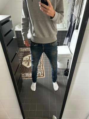 slim fit jeans 29/32 Nudie Jeans - Säljer ett par blå slim fit jeans från Nudie Jeans med snygga slitningar och klassisk femficksmodell. Jeansen har en något tvättad look och är tillverkade i bomull med stretch för skön passform. Perfekta för en avslappnad och trendig stil.