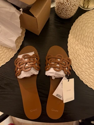 Bruna lädersandaler från Mango - Snygga bruna sandaler från Mango med flätad, dekorativ rem över foten. Platta och stilrena, perfekta för varma dagar. Minimalistisk design med öppen tå och diskret logga på sulan. Storlek 39, box medföljer. ‼️helt oanvänd‼️