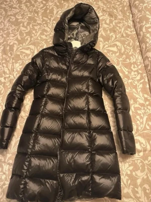 Svart lång dunjacka med huva colmar  - Snygg svart lång dunjacka med huva och quiltad design. Jackan har dragkedja framtill och en diskret logga på ärmen. Tillverkad i glansigt material som ger en modern look. Perfekt för kalla vinterdagar.