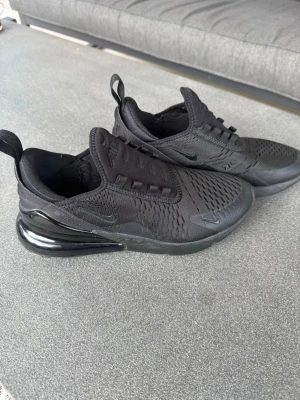 Nike Air Max 270 helsvarta sneakers - Säljer ett par helsvarta Nike Air Max 270 sneakers med mesh-överdel och synlig Air-enhet i hälen. Skorna har en sportig look med diskreta detaljer och dragflikar bak. Perfekta för dig som gillar stilrena och bekväma sneakers.