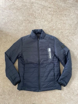 Mörkblå pufferjacka från BOSS - Snygg mörkblå pufferjacka från BOSS med diskret logga på bröstet och patch på ärmen. Jackan har ståkrage, raka ärmar och quiltad design. Dragkedja framtill och lätt vadderad för extra värme. Perfekt för kalla dagar.