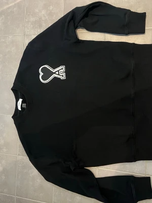 Ami Svart sweatshirt  - Säljer en svart sweatshirt med en stor broderad hjärtdetalj i vitt på bröstet. Tröjan har rund halsringning, långa ärmar och ribbade muddar. Perfekt för dig som gillar stilrena plagg med en twist.