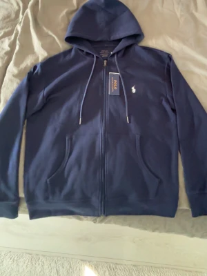 Polo kofta  - Snygg marinblå hoodie från Polo Ralph Lauren med dragkedja framtill och klassisk vit logga på bröstet. Hoodien har två fickor, justerbar huva med snören och är tillverkad i mjukt bomullsmaterial. Perfekt för en avslappnad och stilren look.