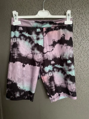 Tie dye cykelshorts i pastell - Säljer ett par trendiga cykelshorts med tie dye-mönster i svart, ljusrosa och ljusblå toner. Shortsen har en midwaist passform och är gjorda i stretchigt syntetmaterial, perfekta för en avslappnad och sportig stil.
