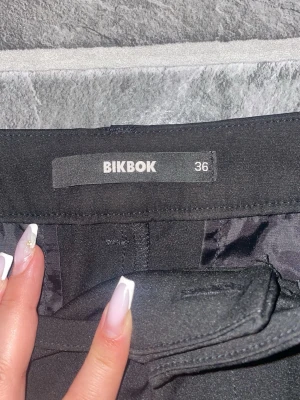 Svarta kostymbyxor short från BikBok lågmidja  - Snygga svarta kostymbyxor från BikBok i storlek 36. Byxorna har raka ben, diskreta fickor och klassisk midja. Perfekta för en stilren look och lätta att matcha med olika toppar. Materialet känns mjukt och följsamt.