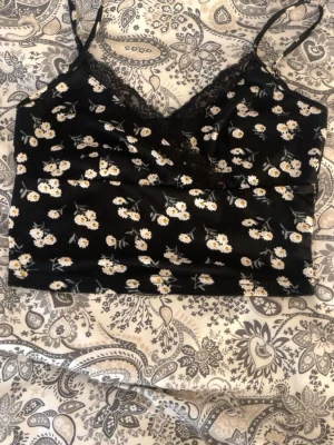 Svart blommig linnetopp med spets - Söt svart linnetopp med vita och gula blommor och tunna axelband. Toppen har spetsdetaljer vid urringningen och är croppad i modellen. Perfekt för sommarens alla tillfällen! Djur finns i hemmet, tvättas innan leverans.🫶🏼