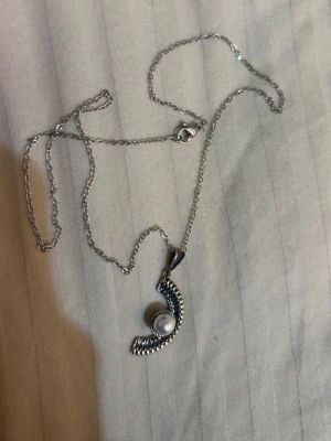 Silverhalsband med pärlhänge - Snyggt halsband i silverfärg med ett unikt böjt hänge dekorerat med små kulor och en vit pärla i mitten. Kedjan är tunn och har ett klassiskt lås. Perfekt accessoar för att lyfta din outfit med en elegant touch.