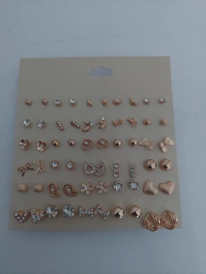 Set med 30 par guldfärgade örhängen - Säljer ett stort set med 30 par örhängen i olika former som hjärtan, blommor, rosetter och stenar. Örhängena är guldfärgade och vissa har gnistrande stenar som liknar pärlor. Perfekt för dig som vill variera din stil med många olika alternativ.