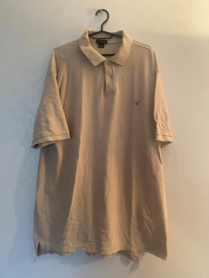 Beige pikétröja från Gant - Klassisk beige pikétröja från Gant med korta ärmar och krage. Tröjan har knappar vid halsen och en liten broderad logga på bröstet. Tillverkad i mjuk bomull för en bekväm känsla.
