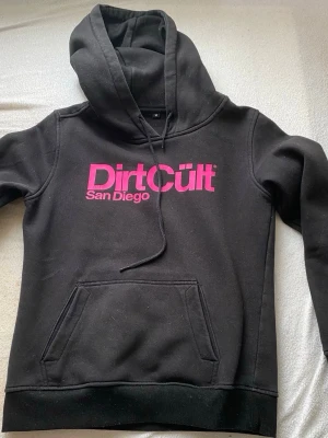 Svart Dirt Cult hoodie med rosa tryck - Snygg svart hoodie från Dirt Cult med stor rosa logga och texten 'San Diego' på bröstet. Klassisk känguruficka framtill och justerbar huva med snören. Perfekt för dig som gillar streetwear och vill sticka ut med färgglatt tryck.
