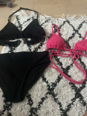 Två bikinis från H&M svart & rosa - Säljer två bikinis från H&M. En svart bikini med volangdetalj på toppen och en rosa ribbad trekantsbikini med knyt i sidan och i nacken. Båda är perfekta för stranden och har en snygg och ungdomlig stil. den svarta är från Lindex 