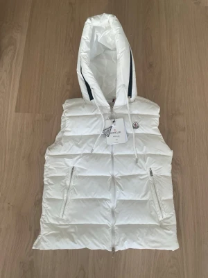 Vit dunväst med huva från Moncler - Snygg vit dunväst från Moncler med stor huva och dragkedja framtill. Västen har två dragkedjeförsedda fickor och Moncler-logga på bröstet. Tillverkad i glansigt material och fodrad med dun för extra värme. Perfekt för lager-på-lager och streetstyle.