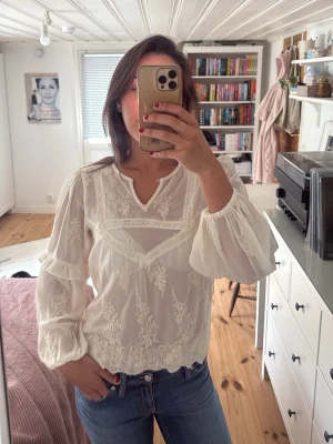 Vit spetsblus med lång ärm - Romantisk vit blus med vackra spetsdetaljer och broderier. Blusen har lång ärm med lätt puff och avslutas med resår vid ärmslut. Rundad halsringning med liten v-skärning framtill. Perfekt att styla med jeans för en trendig look.