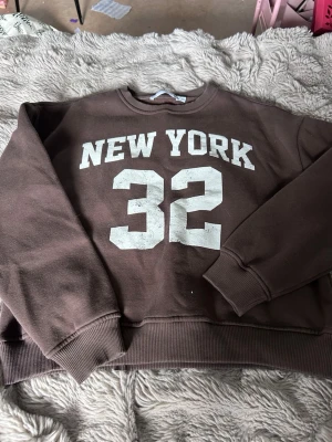 Brun sweatshirt med tryck 'New York 32' - Cool brun sweatshirt med vitt tryck 'NEW YORK 32' på bröstet. Tröjan har rund hals, ribbade muddar vid ärmslut och nederkant samt en avslappnad passform. Perfekt för en sportig och avslappnad stil.