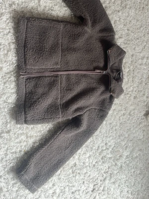 Brun höst jacka från lager 157 - Mysig brun teddyjacka med hög krage och hel dragkedja framtill. Jackan har två stora fickor på framsidan och är gjord i fluffigt teddy-material som håller dig varm. Perfekt för kyliga dagar och enkel att matcha med olika outfits.ser rätt så kort ut men inte på enligt mig.