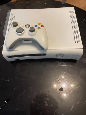 Xbox 360 vit spelkonsol med handkontroll - Säljer en klassisk Xbox 360 i vit färg från Microsoft. Konsolen har en stilren, rektangulär design och är tillverkad i plast. Medföljer gör en matchande vit trådlös handkontroll och strömadapter. Perfekt för gaming och retrospel!