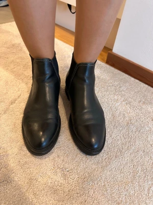 Svarta Chelsea boots från Gant - Säljer ett par klassiska svarta Chelsea boots från Gant i slätt läder. Bootsens design är stilren med elastiska paneler på sidorna och rundad tå. Gant-logga finns diskret på sidan. Perfekta för en clean och tidlös look.
