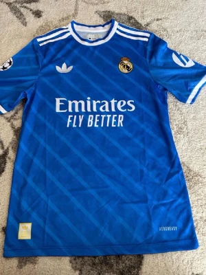 Blå Real Madrid matchtröja Adidas - Snygg blå Real Madrid matchtröja från Adidas med vita detaljer och tryck. Framsidan har klubbmärke, Adidas-logga och Emirates Fly Better-tryck. Baksidan har Mbappé och nummer 10. Tillverkad i lätt funktionsmaterial (AEROREADY), perfekt för fotboll.