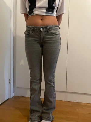 Grå bootcut jeans med fickdetaljer - Säljer ett par grå bootcut jeans med låg midja och snygga fickdetaljer bak med lock och knapp. Jeansen har klassisk femficksmodell och är i stretchigt denimtyg som sitter tajt över låren och släpper ut vid bensluten.
