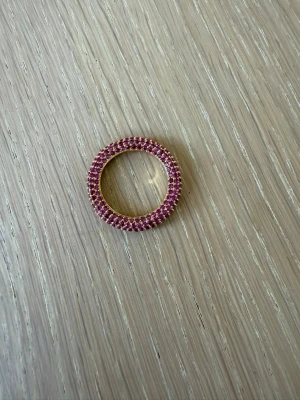 Guldfärgad ring med rosa stenar - Snygg guldfärgad ring täckt med små rosa stenar runt hela bandet. Ringen har en rund form och gnistrar fint i ljuset. Perfekt för dig som vill ha en färgklick på fingret och gillar detaljer som sticker ut.