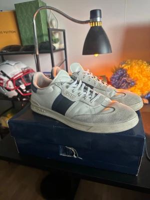 Vita Polo Ralph Lauren sneakers - Snygga vita sneakers från Polo Ralph Lauren med marinblå detalj på sidan och logga. Skorna har lågt skaft, vita skosnören och ovandel i mocka och läder. Perfekta för dig som gillar klassisk och stilren streetwear. Finns kvitto om ni är intresserade 