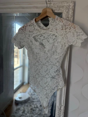 Vit spetsbody med kort ärm - Säljer en superfin vit body i spets med korta ärmar och rund hals. Bodyn har ett vackert blommigt spetsmönster och är lätt transparent. Perfekt för dig som vill ha något extra snyggt under kläderna eller till en cool outfit.