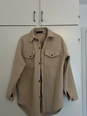 Beige overshirt med fickor - Stilren beige overshirt med knappar framtill och två stora bröstfickor med lock. Skjortan har klassisk krage, långa ärmar med knapp vid ärmslut och rundad nederkant. Perfekt lager-på-lager-plagg i mjukt material.