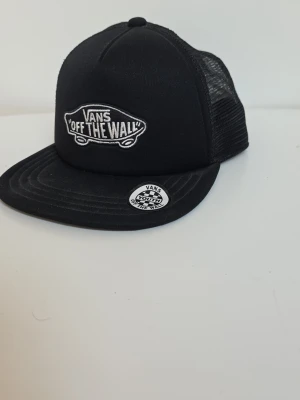 Svart truckerkeps från Vans - Säljer en svart truckerkeps från Vans med klassisk 'Off The Wall'-logga framtill. Kepsen har böjd skärm och mesh på sidorna och bak för extra ventilation. Justerbar baktill för perfekt passform. Perfekt för dig som gillar streetwear och skatekultur. Använts 1 gång