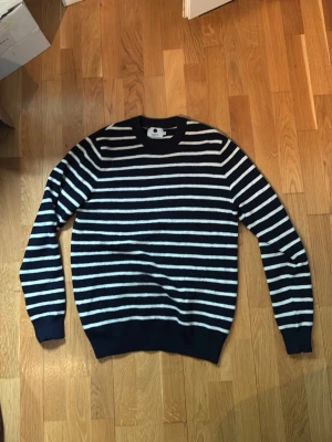 Marinblå randig tröja från NN07 - Snygg marinblå stickad tröja från NN07 med vita horisontella ränder. Tröjan har rund halsringning och ribbade muddar vid ärmslut och nederkant. Perfekt för dig som gillar klassisk och stilren design. Är i storlek S men kan passa M. (Pris kan diskuteras)