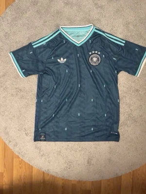 Tysklands retro fotbollströja Adidas - Mörkblå retro fotbollströja från Adidas med Tysklands landslagsmärke och fyra stjärnor på bröstet. Tröjan har ljusblå detaljer, klassiska Adidas-ränder på axlarna och V-ringad krage. Tillverkad i lätt och ventilerande material, perfekt för träning eller match.