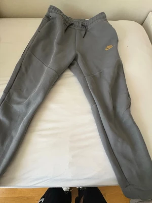 Gråa mjukisbyxor från Nike - Säljer ett par gråa mjukisbyxor från Nike med gul logga på vänster sida. Byxorna har dragsko i midjan, ribbade muddar vid bensluten och praktiska fickor. Tillverkade i mjukt och bekvämt material, perfekta för chill dagar.