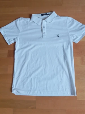Vit pikétröja från Ralph Lauren - Klassisk vit pikétröja från Ralph Lauren med korta ärmar och krage. Tröjan har knappar framtill och den ikoniska broderade loggan på bröstet. Tillverkad i bomull för en bekväm och stilren look.