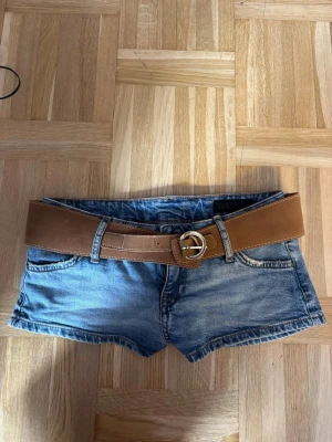 Blå jeansshorts med brunt bälte - Säljer ett par blå denimshorts med ett brett brunt bälte. Shortsen har klassisk femficksdesign och är korta i modellen. Perfekta för varma dagar och ger en avslappnad look. Bältet har ett snyggt metallspänne som detalj. Använda en gång.