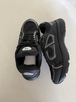 Svarta Dior sneakers med vita detaljer - Snygga svarta sneakers från Dior med vita och grå detaljer. Skorna har en chunky sula, meshpaneler och reflekterande linjer på sidan. Klassisk snörning och logga på plösen samt insidan. Perfekta för dig som vill ha en exklusiv och trendig look.