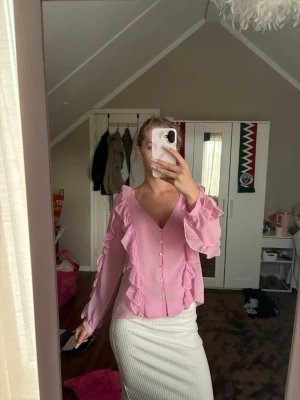 Rosa blus med volanger och pärlknappar - Supersöt rosa blus med långa ärmar, volangdetaljer längs fram- och baksidan samt pärlknappar framtill. Blusen är lätt transparent och har en djup v-ringning. Perfekt för dig som vill ha en feminin och trendig look.